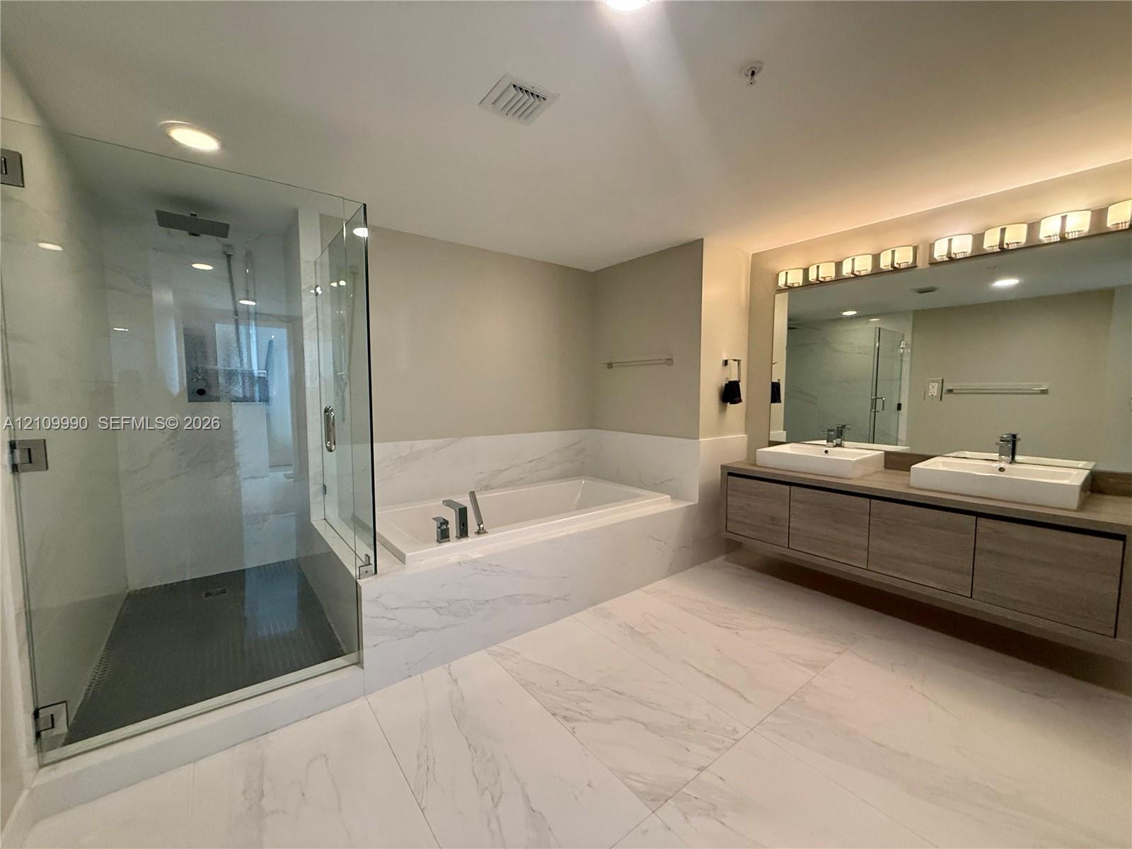 Bathroom featuring a glass-enclosed shower, soaking tub with stone-pattern surround, dual-sink floating vanity with wood-finish cabinetry, and large-format floor tiling