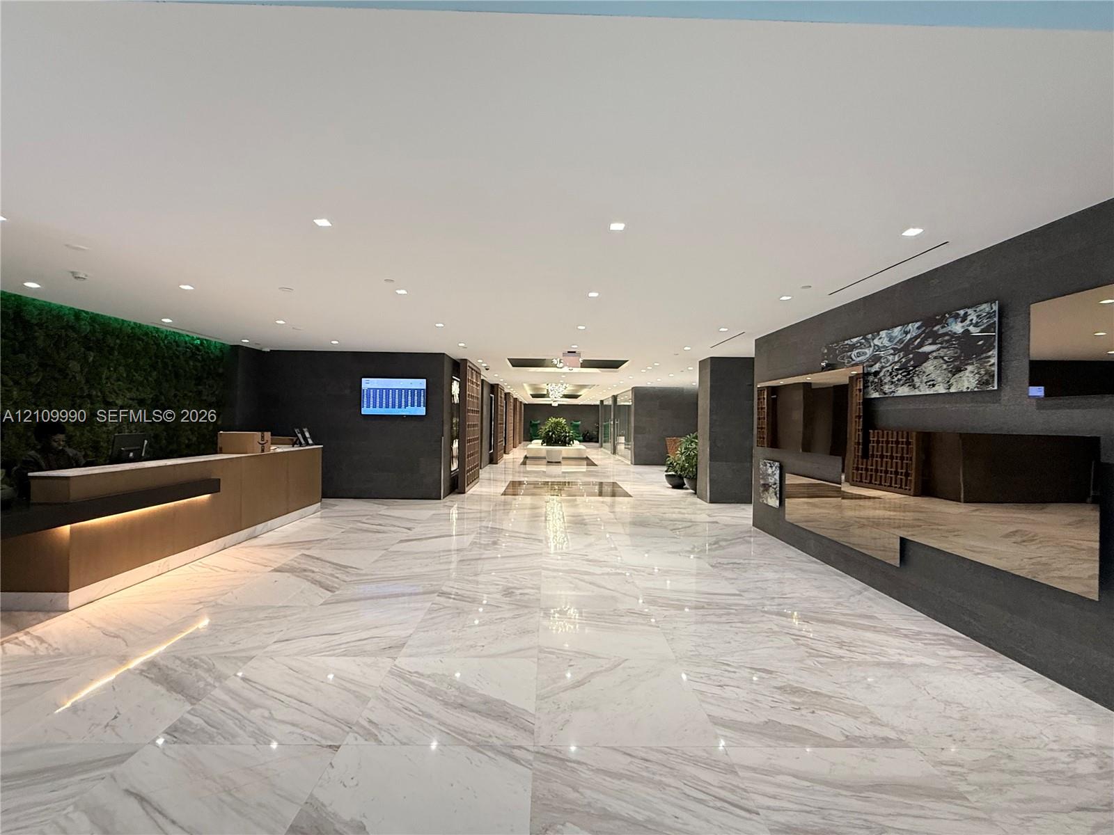 Lobby interior featuring large-format polished stone flooring, a built-in reception desk with integrated under-counter lighting, a full-height living wall installation, recessed ceiling lighting, and dark stone-finish accent walls