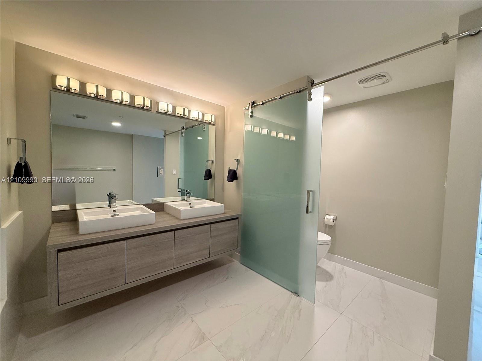 Floating wood-finish vanity with dual rectangular vessel sinks, expansive wall-to-wall mirror, oversized multi-bulb light fixture, and frosted glass sliding door