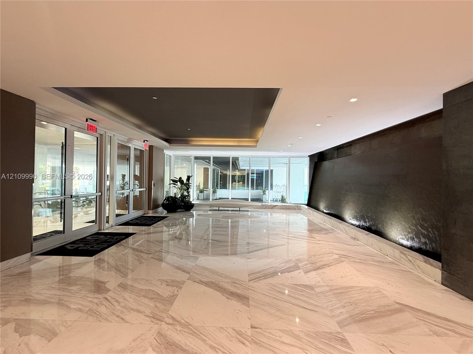 Spacious lobby featuring polished stone flooring in a geometric pattern, an expansive dark-toned accent water wall, recessed ceiling detailing, and glass-paneled entry doors