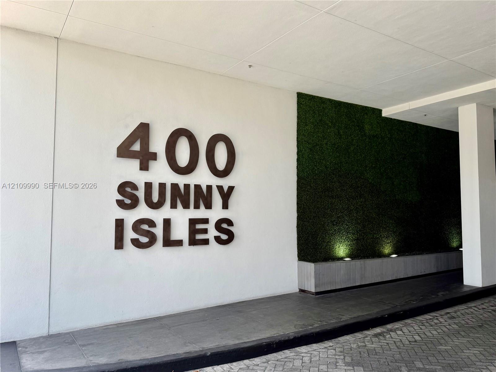 Covered entryway featuring dimensional signage, a green wall accent, concrete planter base, and stone paver driveway