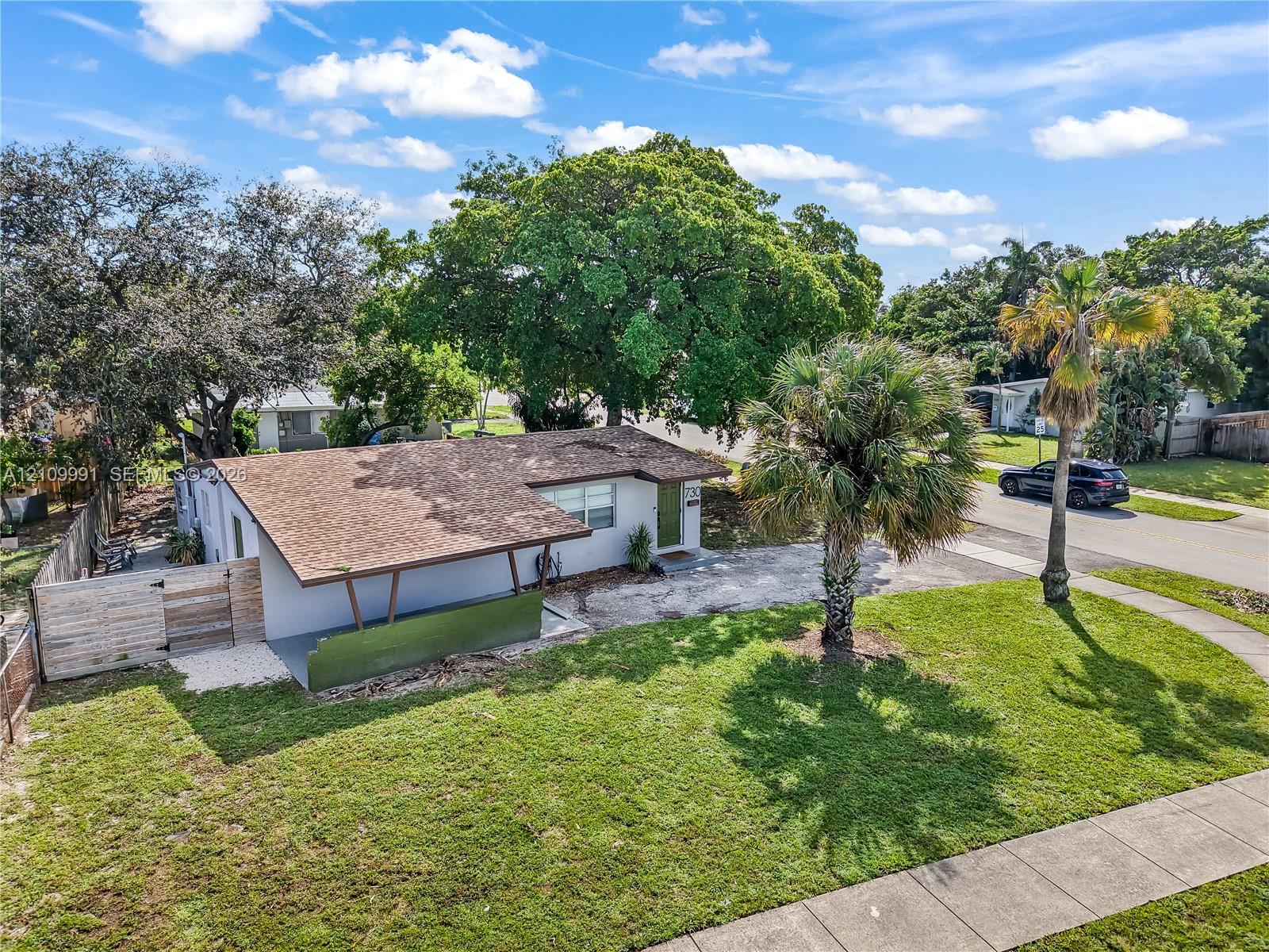 Single-story exterior featuring a shingled roof, overhanging carport, integrated wood-finish fencing, and established palm and shade trees Single-story exterior featuring a shingled roof, overhanging carport, integrated wood-finish fencing, and established palm and shade trees