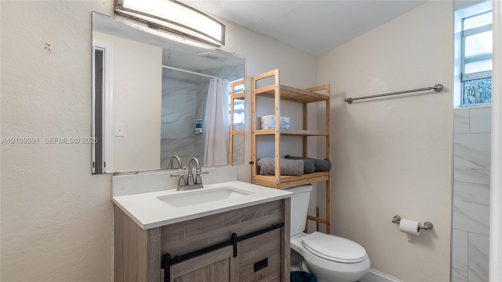 Bathroom featuring a wood-finish vanity with a stone-composite countertop, undermount sink, dual-handle faucet, large wall-mounted mirror with integrated lighting, and a shower area with light-toned tiled walls