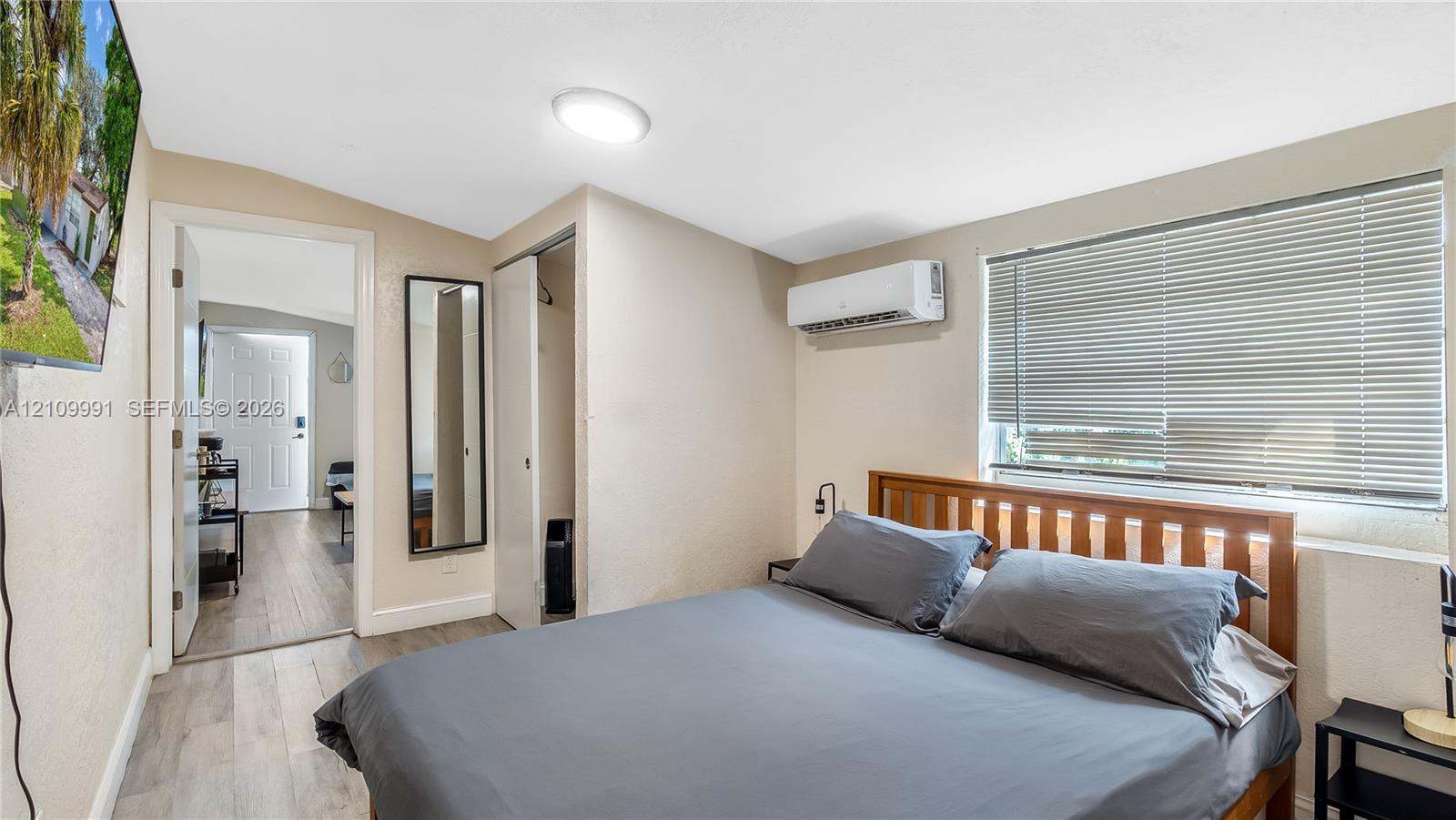 Neutral-toned room featuring wood-finish flooring, a wall-mounted ductless air conditioning unit, and a window with horizontal blinds