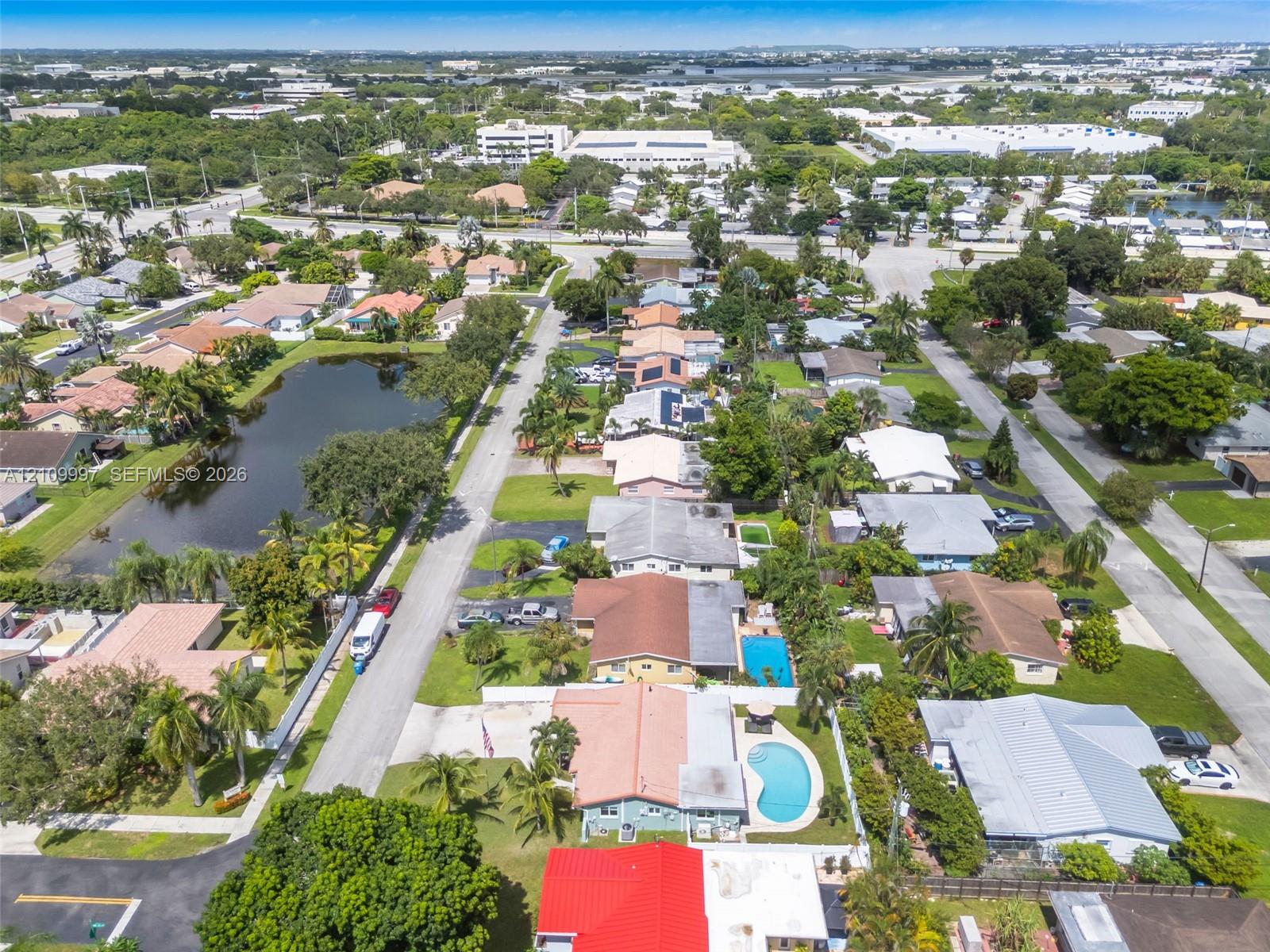 Aerial perspective of a residential neighborhood featuring paved roadways, established landscaping with mature palm trees, single-story residences with diverse rooflines, and localized water features