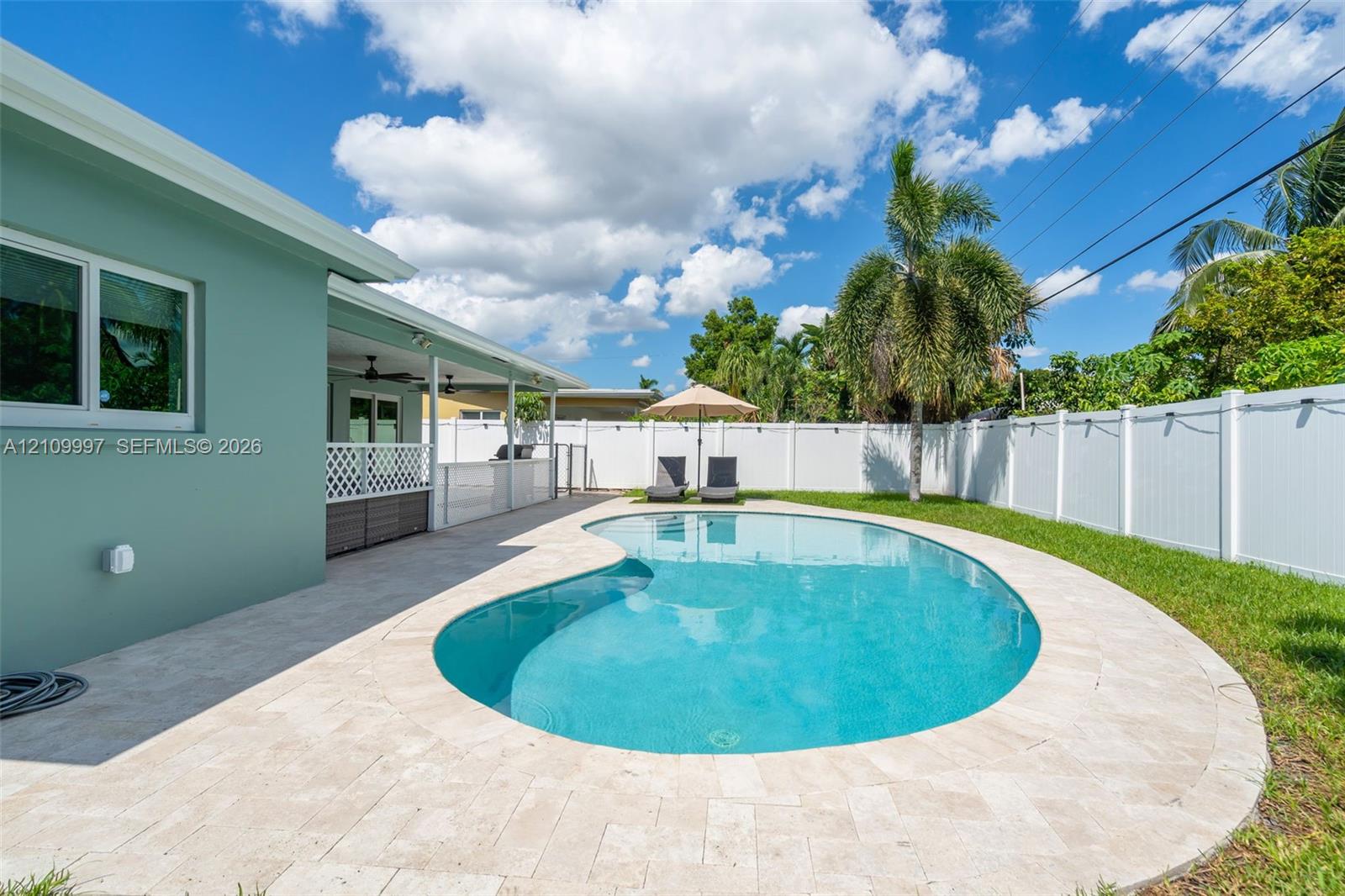 In-ground pool featuring a curved design and light-toned stone paver decking, enclosed by a white vinyl privacy fence and a covered patio area