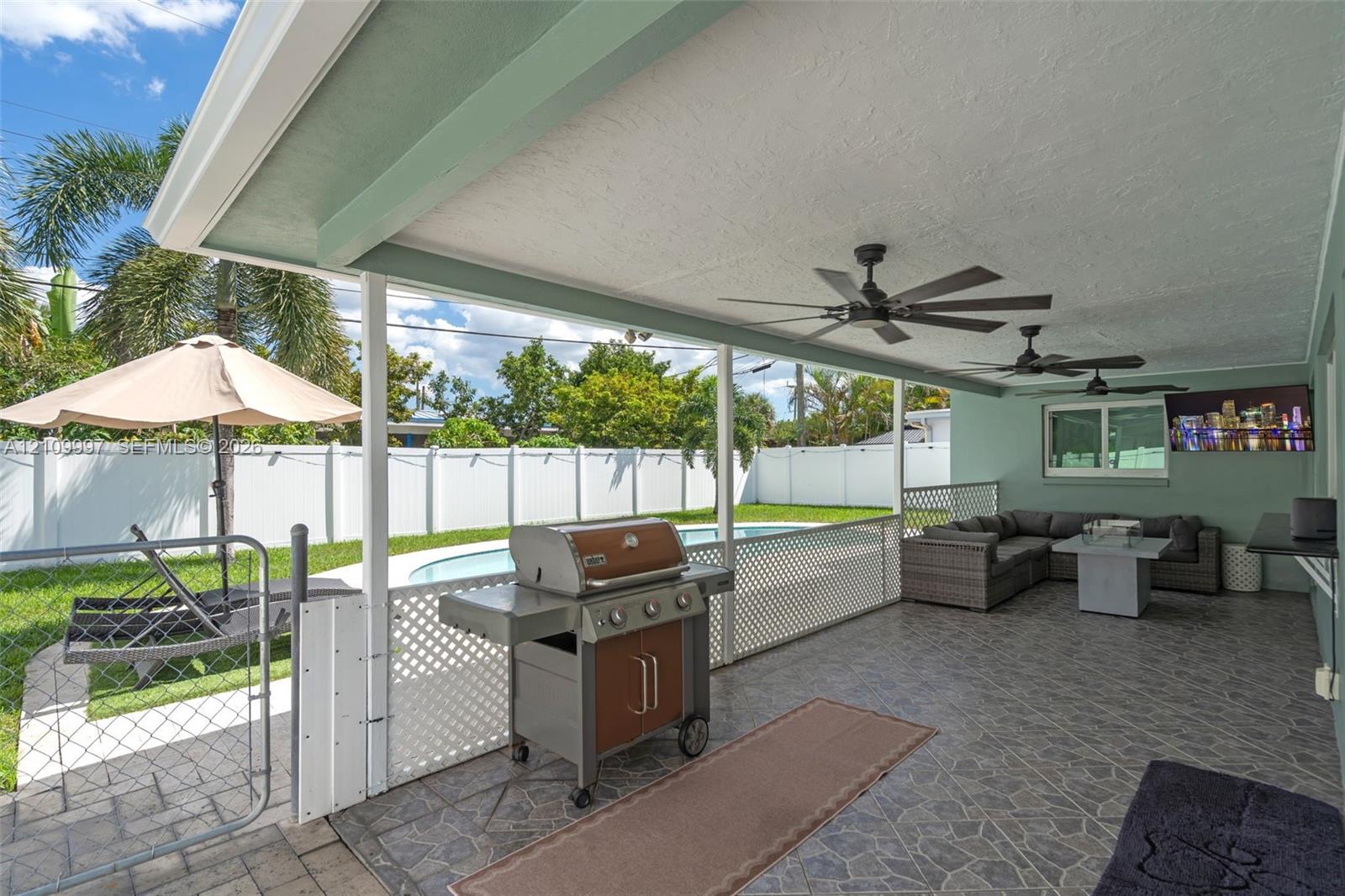 Covered patio featuring stone-patterned tile flooring, recessed ceiling fans, and a white lattice privacy screen, opening to an outdoor area with a swimming pool and vinyl perimeter fencing