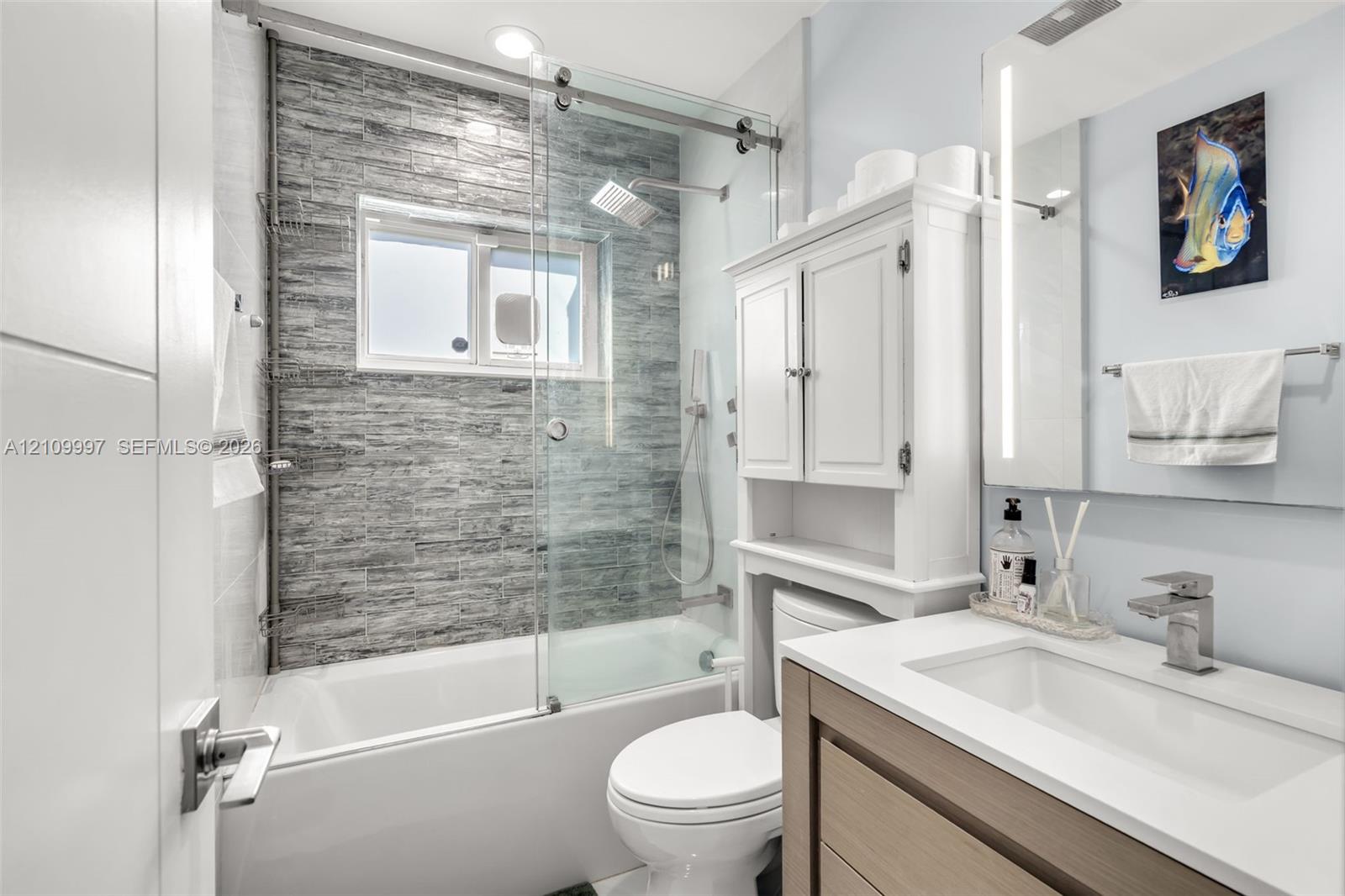 Bathroom featuring a shower-tub combination with a glass sliding door, textured stone-tile wall, built-in cabinetry, integrated vanity lighting, and a square vessel sink
