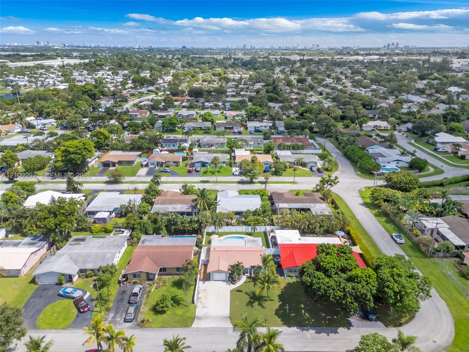 Aerial perspective featuring a suburban residential lot with a concrete driveway, a private in-ground swimming pool, a multi-pitched roof, and mature perimeter trees