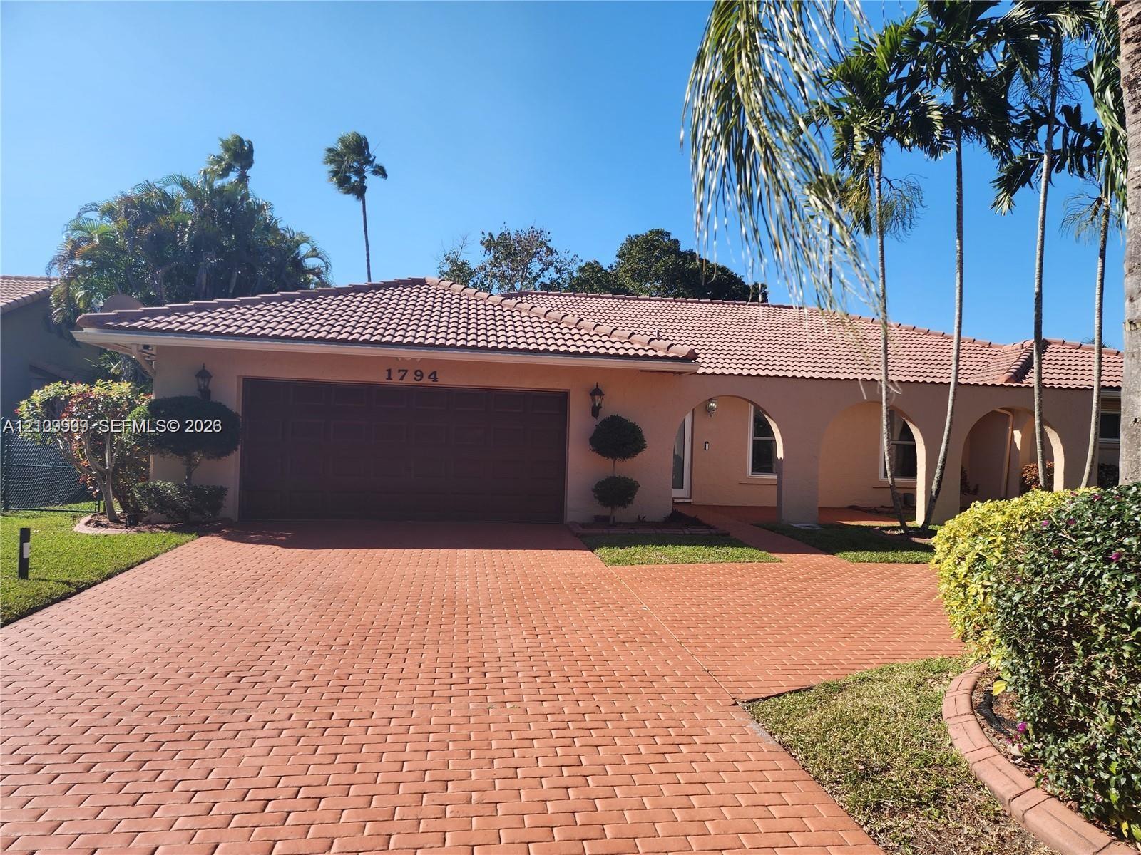 Stucco exterior featuring a clay tile roof, arched covered entry, paver driveway, and mature landscaping