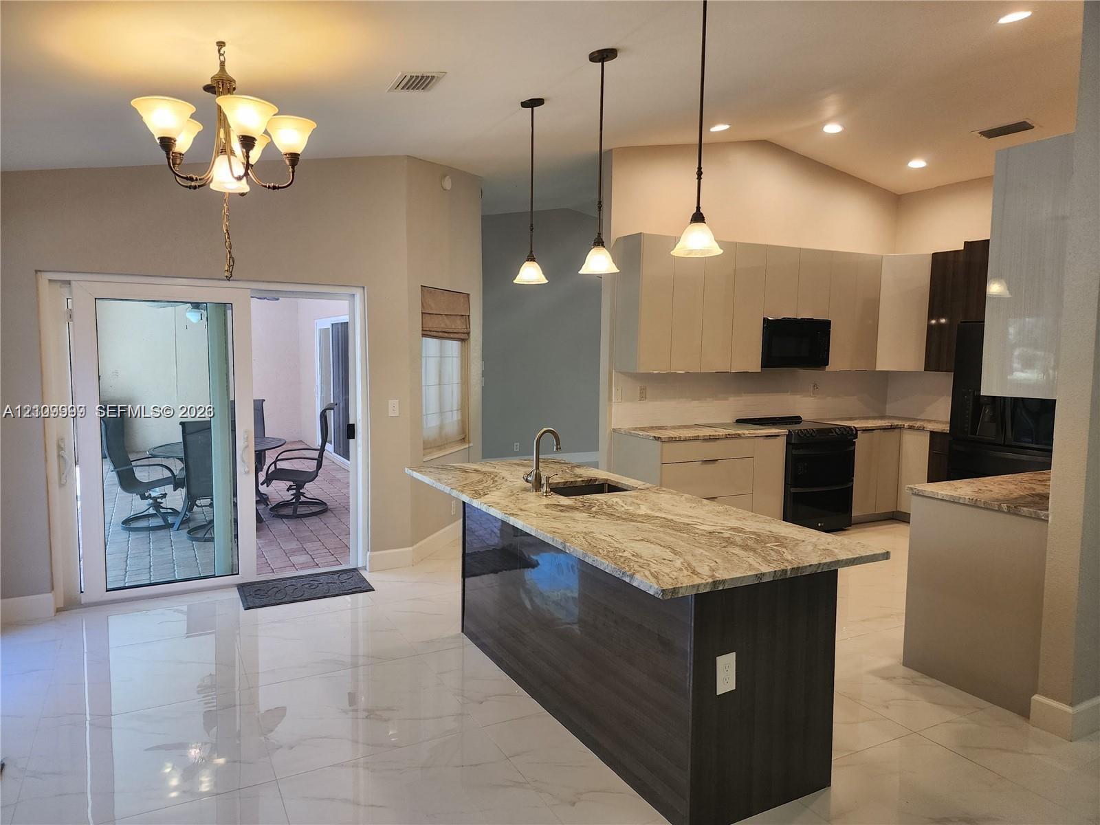Kitchen featuring stone-topped island with undermount sink, flat-panel cabinetry, porcelain tile flooring, trio of pendant lights, and sliding glass doors