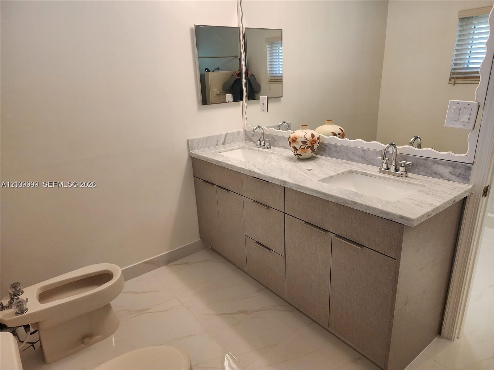 Dual-sink vanity with stone countertop, polished chrome hardware, large vanity mirror, and light-toned tile flooring