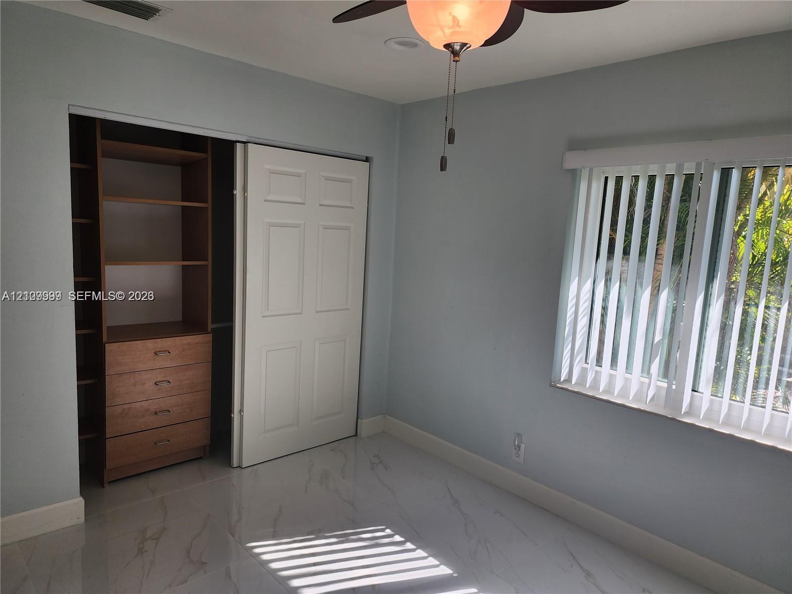 Room featuring large-format tile flooring, a custom-built wood closet system with integrated shelving and drawers, a ceiling fan with integrated lighting, and vertical window blinds