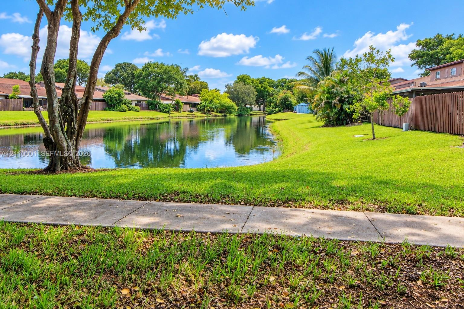 Waterfront grounds featuring a concrete walking path, mature trees, manicured lawn, and perimeter wood fencing