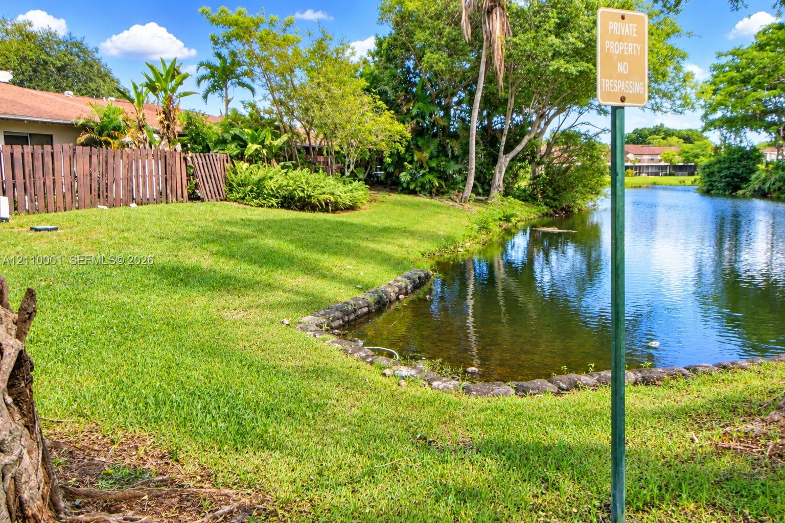 Waterfront lot featuring a natural grass lawn, stone-lined bulkhead, mature trees, and wooden perimeter fencing