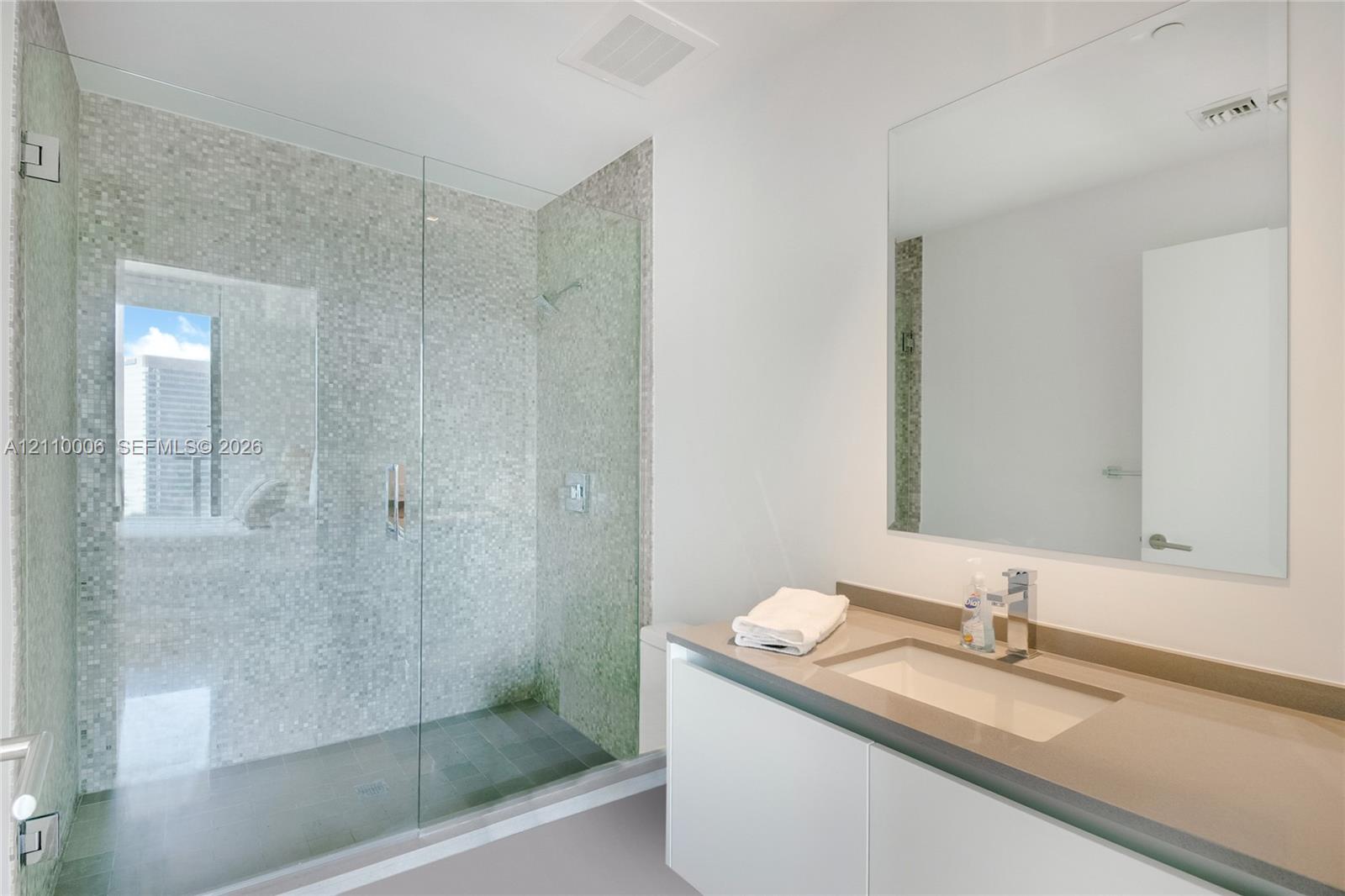 Walk-in shower featuring a glass enclosure and metallic mosaic tile surround, complemented by a stone-surface vanity with an integrated rectangular sink and wall-mounted mirror
