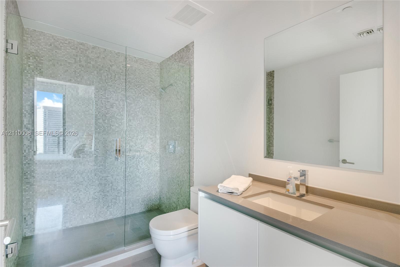 Walk-in shower featuring a glass enclosure and metallic mosaic tile surround, white wall-mounted vanity, rectangular undermount sink, solid surface countertop, and large wall-mounted mirror
