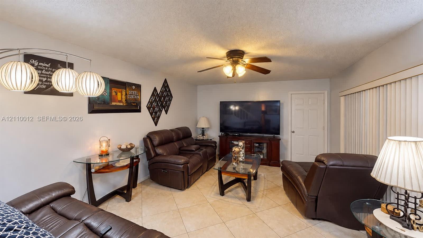 Living area featuring diagonal-set tile flooring, a central ceiling fan with integrated lighting, and vertical window blinds