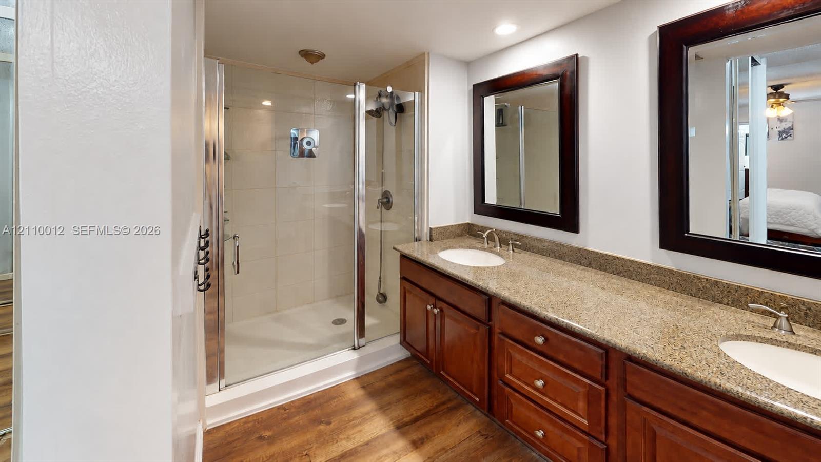 Dual-sink vanity with stone-finish countertop and wood-finish cabinetry, frameless glass shower enclosure with tiled walls, and wood-finish flooring