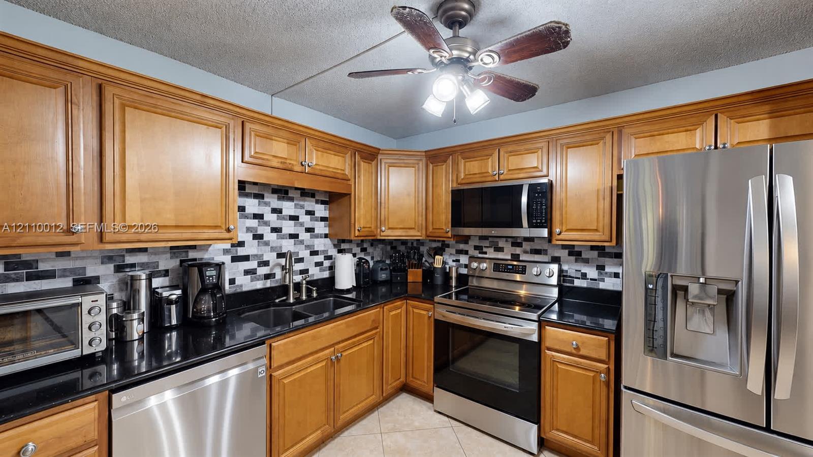 Kitchen featuring natural finish wood cabinetry, contrasting black countertops, a multi-tonal rectangular tile backsplash, stainless steel appliances, and a ceiling-mounted fan with integrated lighting