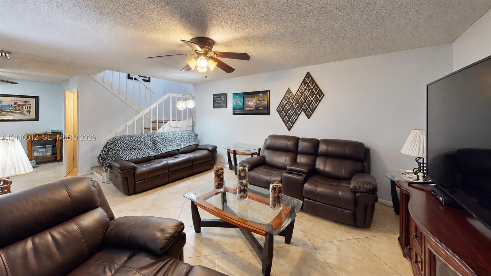 Living area featuring large-format tile flooring, a ceiling fan with integrated lighting, and a white staircase with a metal railing