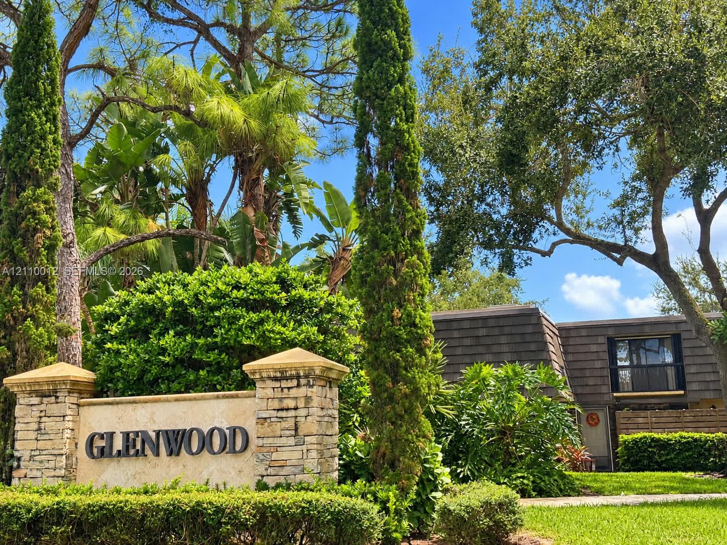 Stone masonry monument sign with capped columns, mature evergreen trees, lush shrubbery, and palm landscaping
