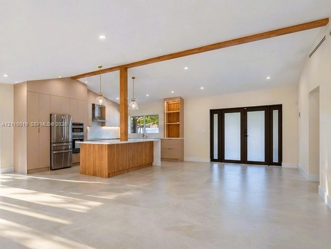 Open-concept interior featuring vaulted ceilings with exposed wood beams, large-format tile flooring, a kitchen island with wood-finish cabinetry, built-in floor-to-ceiling cabinetry, and French doors