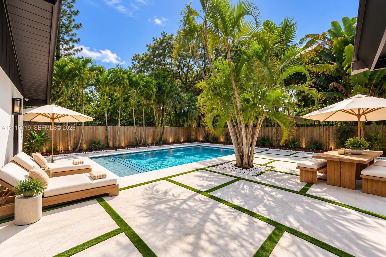 In-ground swimming pool with steps, surrounded by light-toned rectangular stone pavers and synthetic turf inlay, enclosed by a wood-slat perimeter fence and mature palm trees