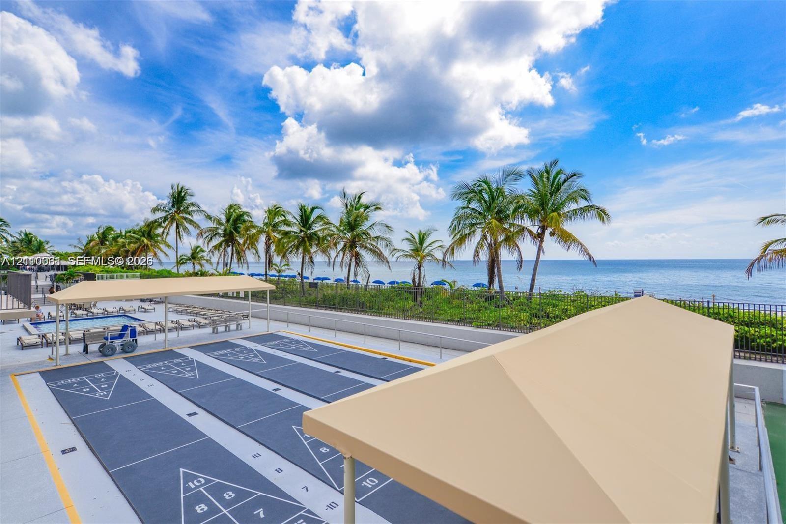 Outdoor recreation area featuring multiple shuffleboard courts, shaded canopies, a swimming pool, and direct oceanfront positioning