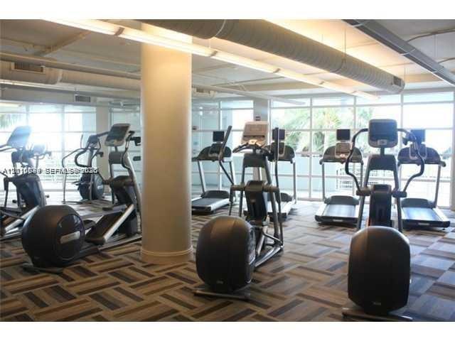 Fitness center featuring floor-to-ceiling windows, patterned modular carpeting, exposed structural support columns, and industrial-style overhead lighting