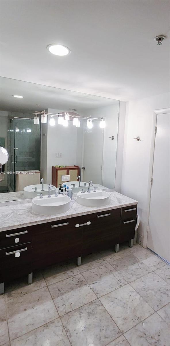 Double vanity featuring dark wood-finish cabinetry, a stone-style countertop, dual round drop-in sinks, and a large-format mirror with integrated overhead lighting