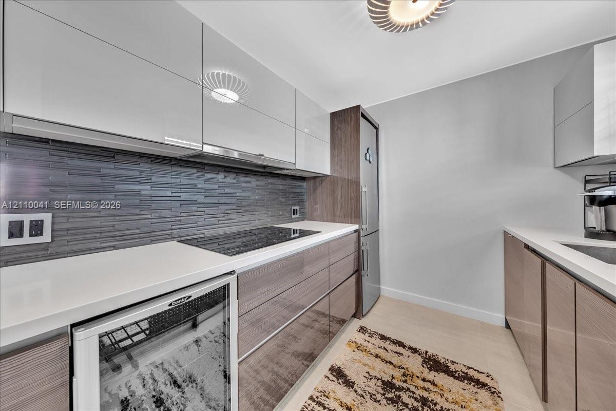 Modern kitchen featuring high-gloss cabinetry, linear tile backsplash, solid-surface countertops, integrated cooktop, and built-in beverage refrigerator