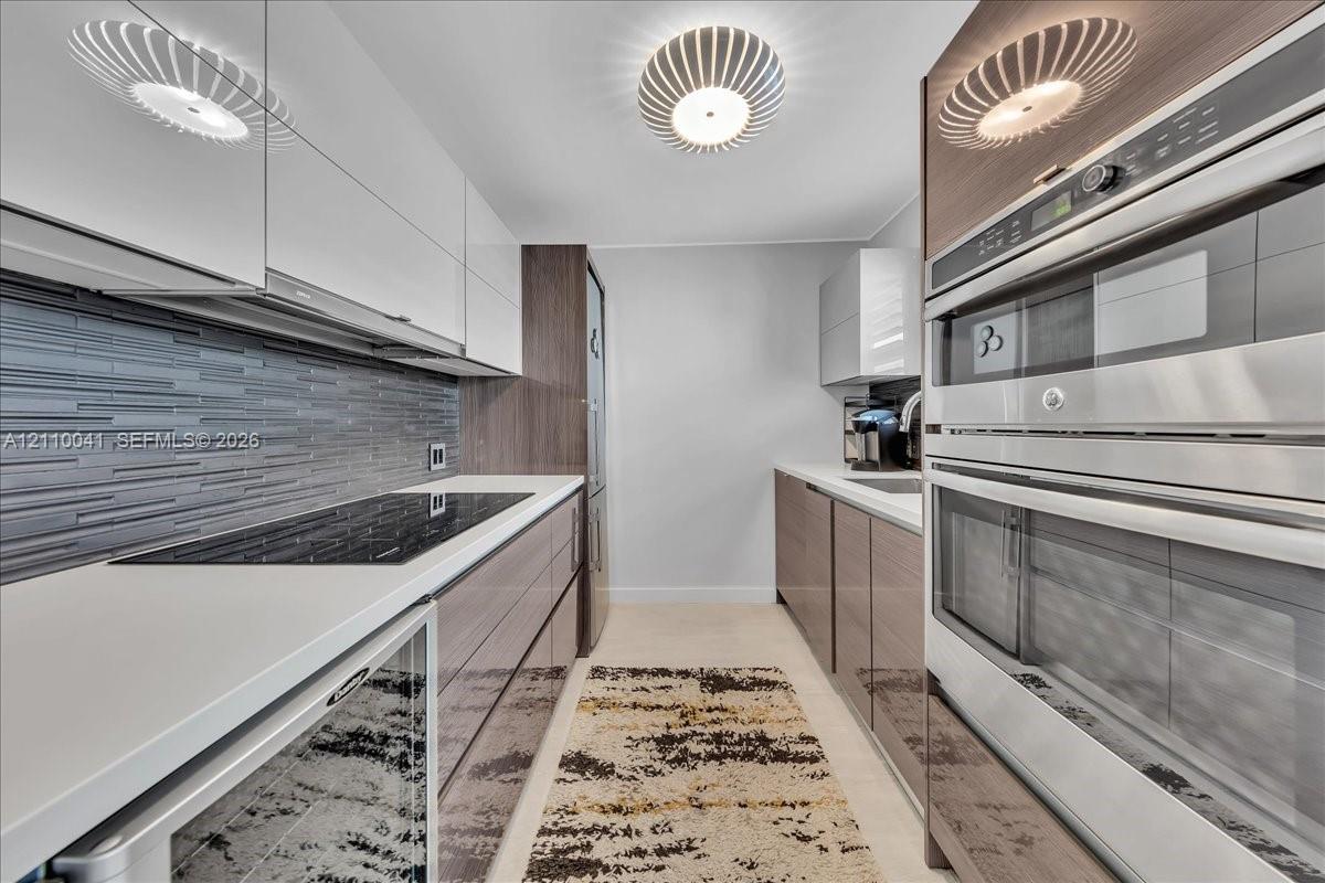 Contemporary kitchen featuring high-gloss cabinetry, stainless steel double wall ovens, a glass cooktop, stone-finish countertops, and a textured tile backsplash
