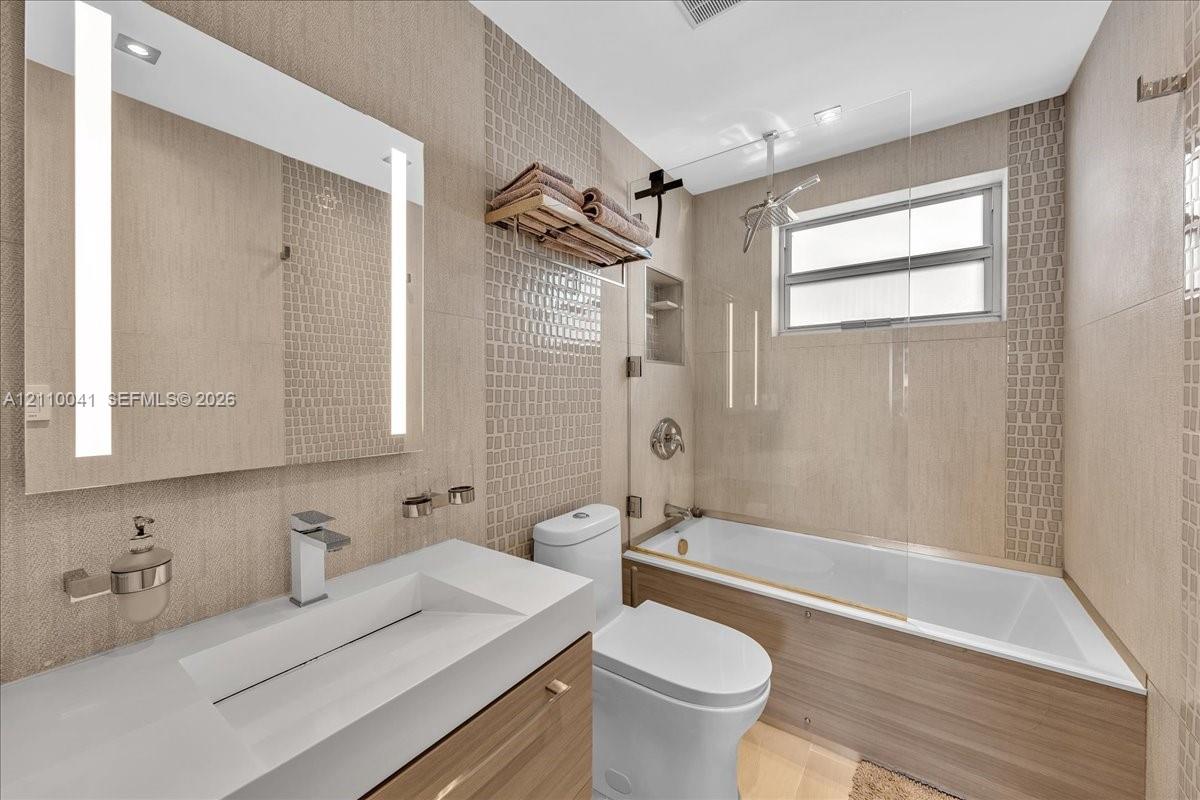 Bathroom featuring a wood-finish vanity with a white rectangular vessel sink, a wall-mounted LED-backlit mirror, a glass-enclosed shower and tub combination, and neutral textured tile wall accents
