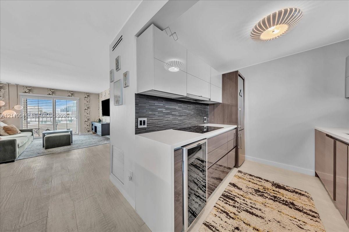 Kitchen featuring high-gloss cabinetry, stone-surface countertops, a textured tile backsplash, integrated glass-door beverage chiller, and a sculptural flush-mount ceiling fixture