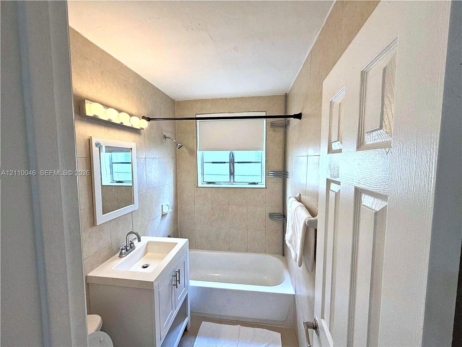 Bathroom featuring ceramic tile wall surround, white vanity cabinet with integrated sink, rectangular mirror, multi-bulb vanity light, and a built-in shower-tub combination