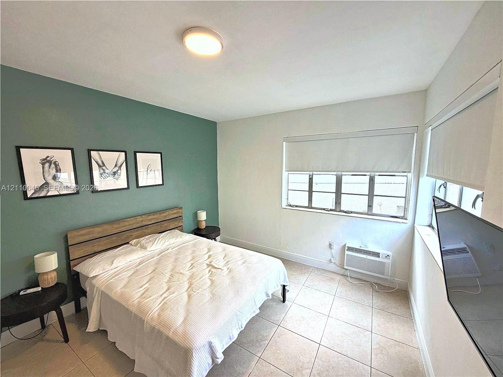 Neutral-tone tiled flooring, accent wall in muted sage, multi-pane window with installed roller shade, and wall-mounted air conditioning unit