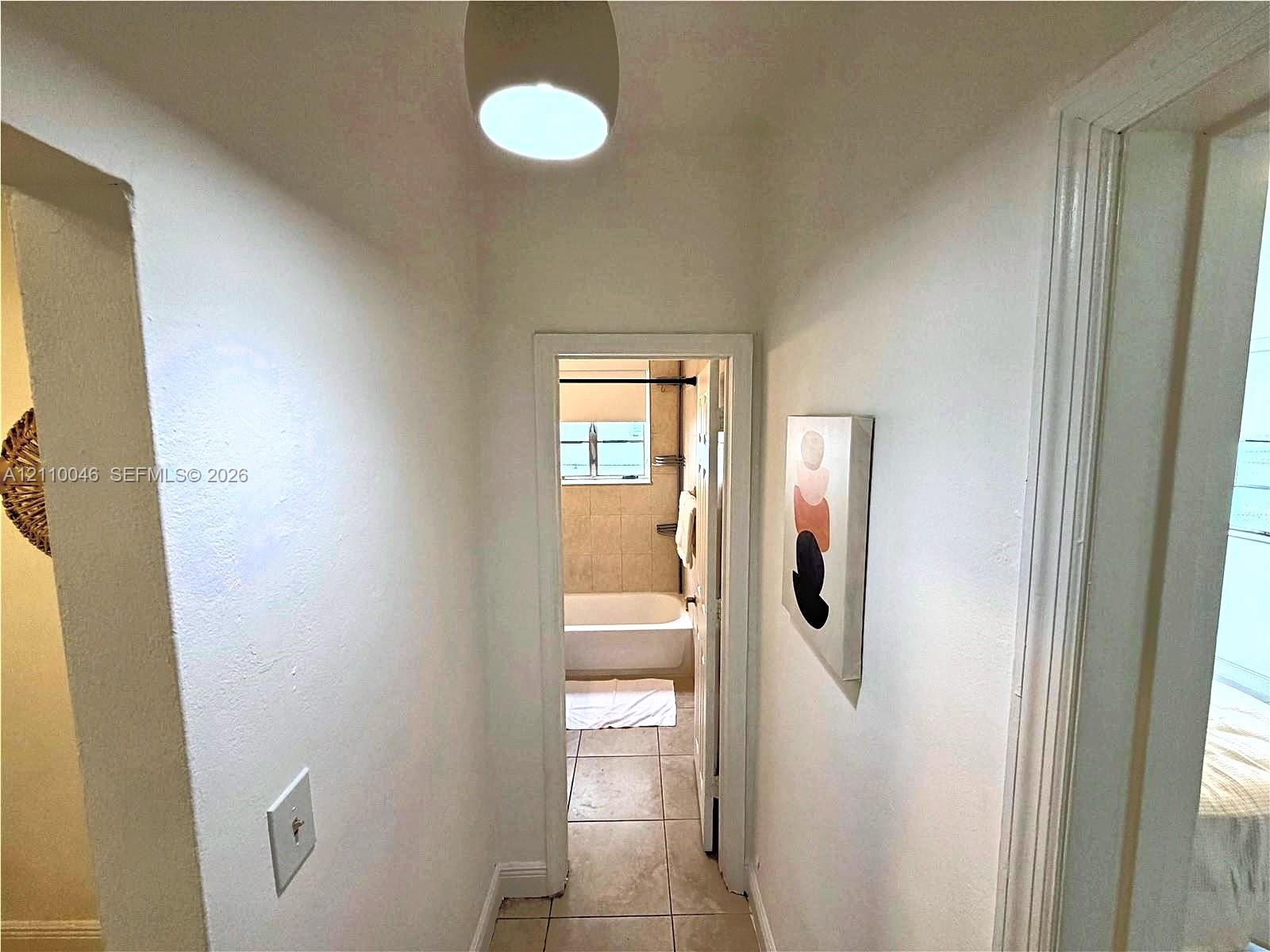 Hallway featuring neutral-toned walls, tile flooring, a semi-flush mount light fixture, and a doorway opening into a bathroom containing a porcelain tub and tile surround