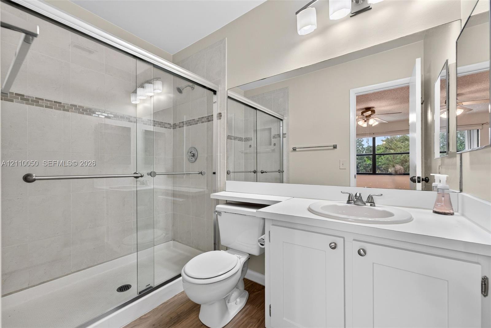 Bathroom featuring a glass-enclosed shower with tiled surround, white cabinetry, integrated sink, wood-finish flooring, and a full-width mirror