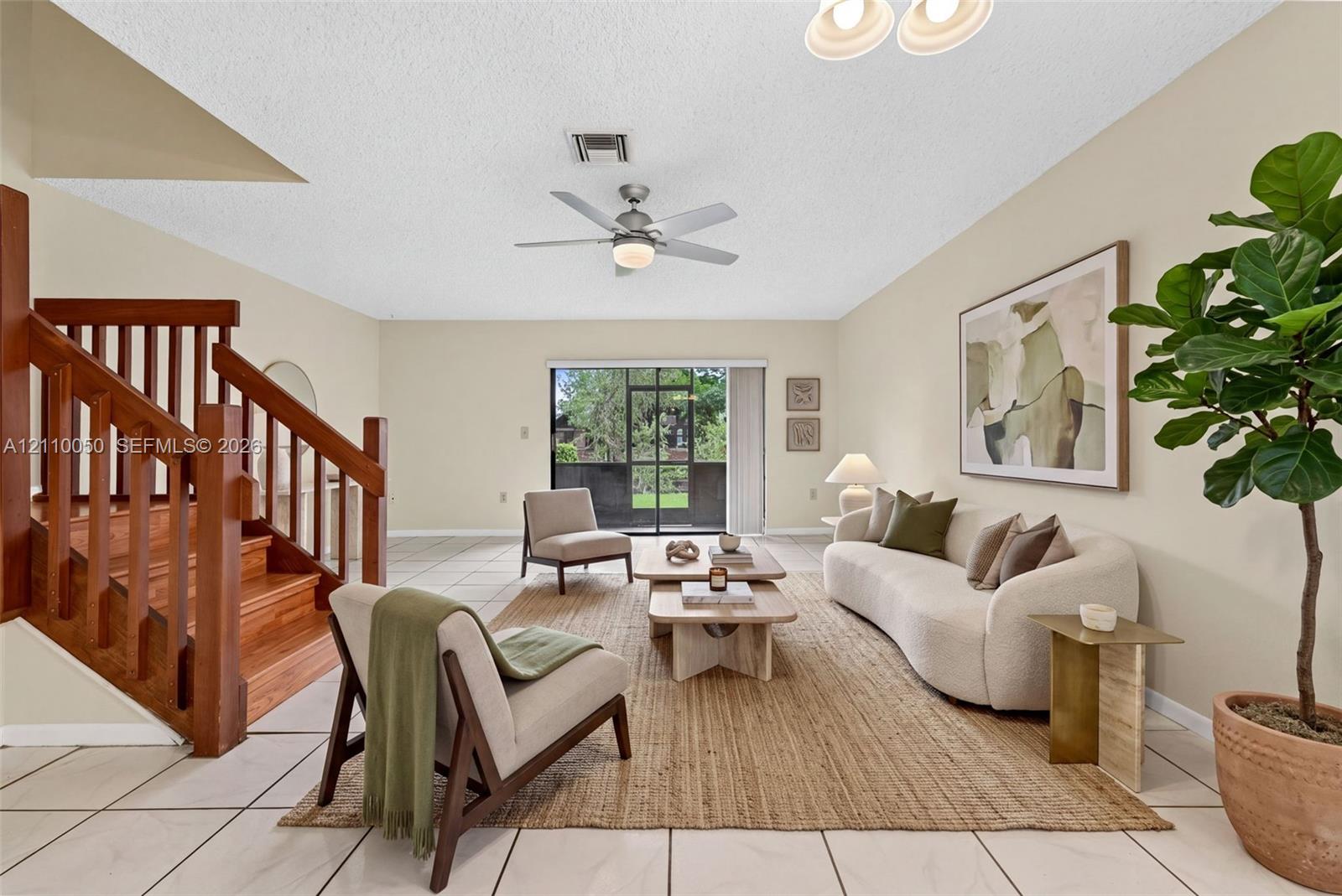 Living area featuring large-format tile flooring, wood-finished staircase with matching balusters, central ceiling fan, and sliding glass doors with a screened enclosure