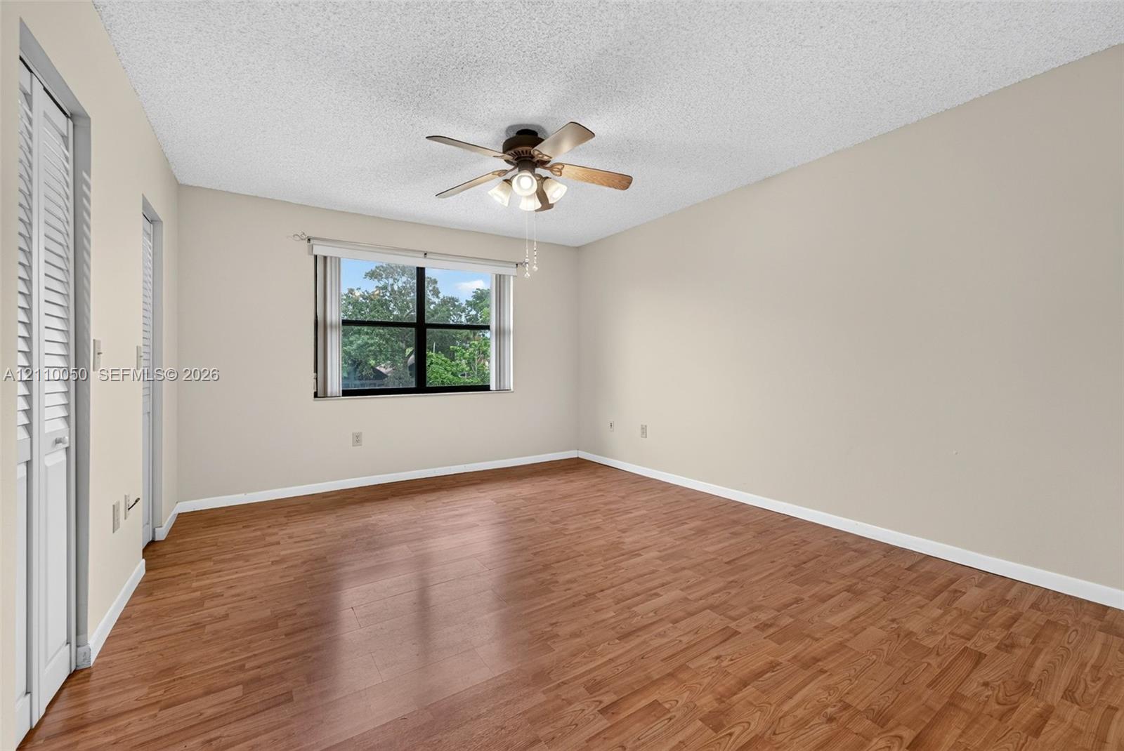 Neutral-toned room featuring wood-finish flooring, a ceiling fan with integrated lighting, louvered closet doors, and a window with exterior greenery
