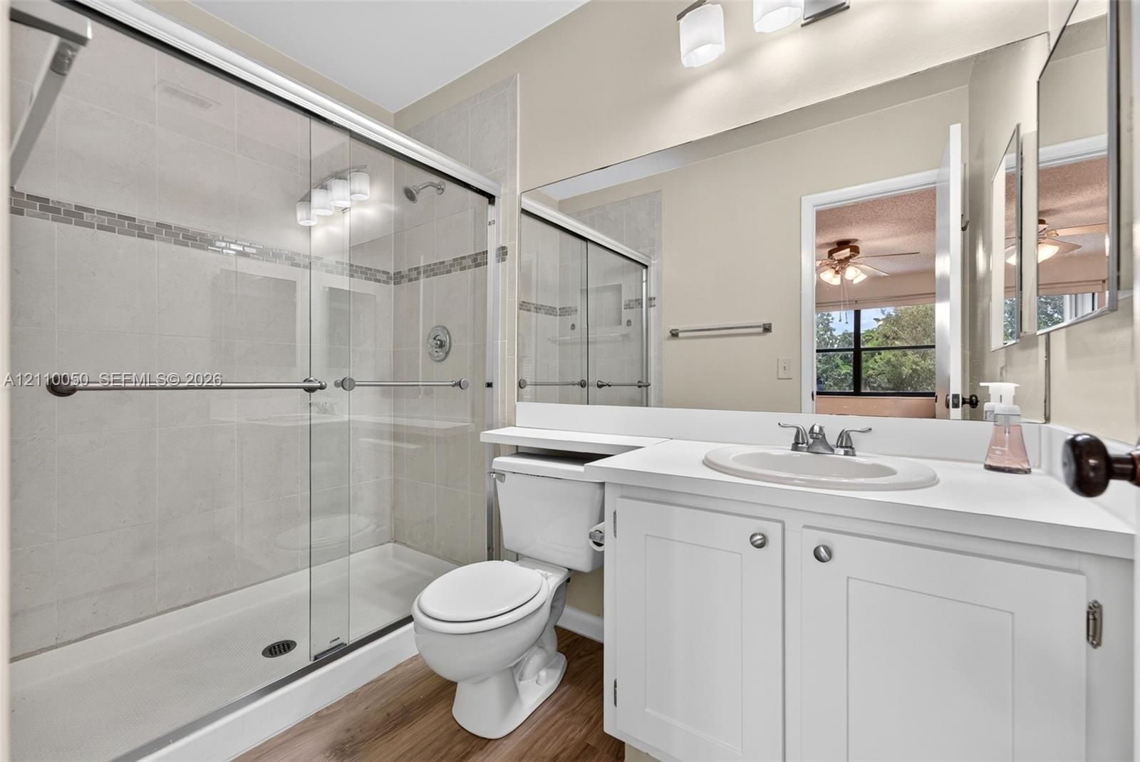 Full-width vanity with an integrated sink, glass-enclosed shower with mosaic tile inlay, wood-finish flooring, and wall-to-wall mirror