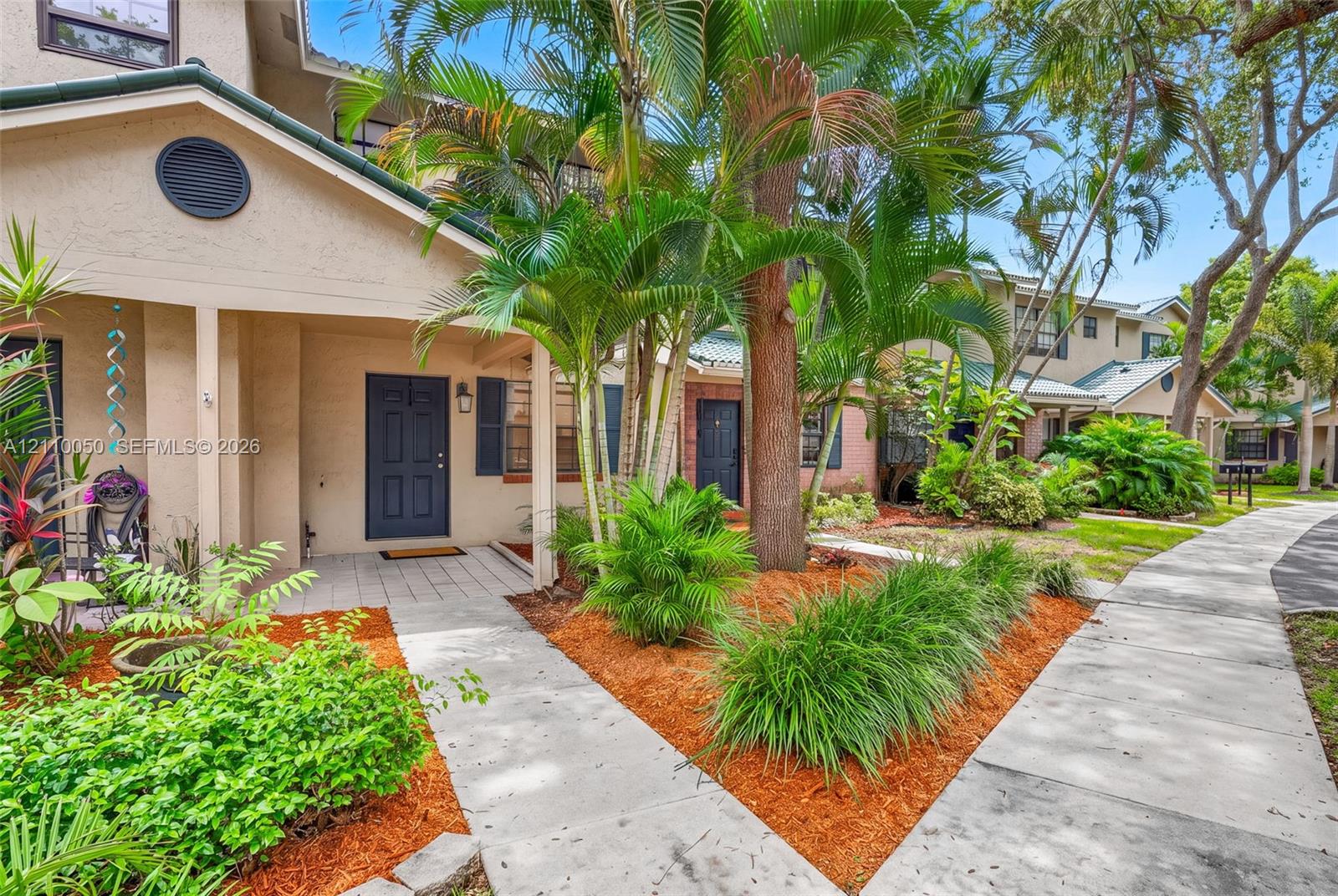 Exterior facade featuring a stucco finish, covered entryway, panel-style front door, paved walkway, and mature tropical landscaping