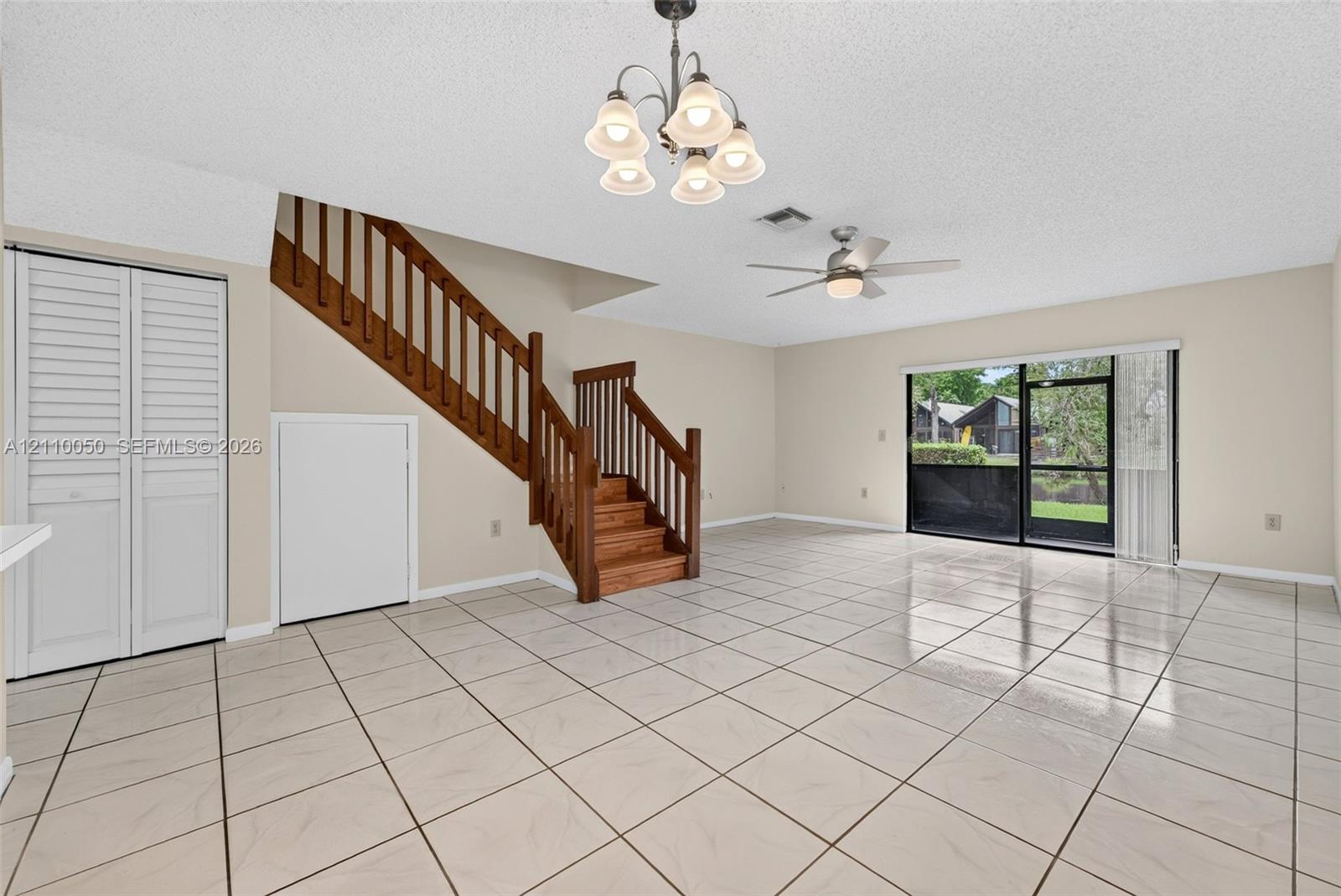 Open-concept living space featuring ceramic tile flooring, a wooden staircase with integrated railing, sliding glass doors, a multi-arm chandelier, and a ceiling fan
