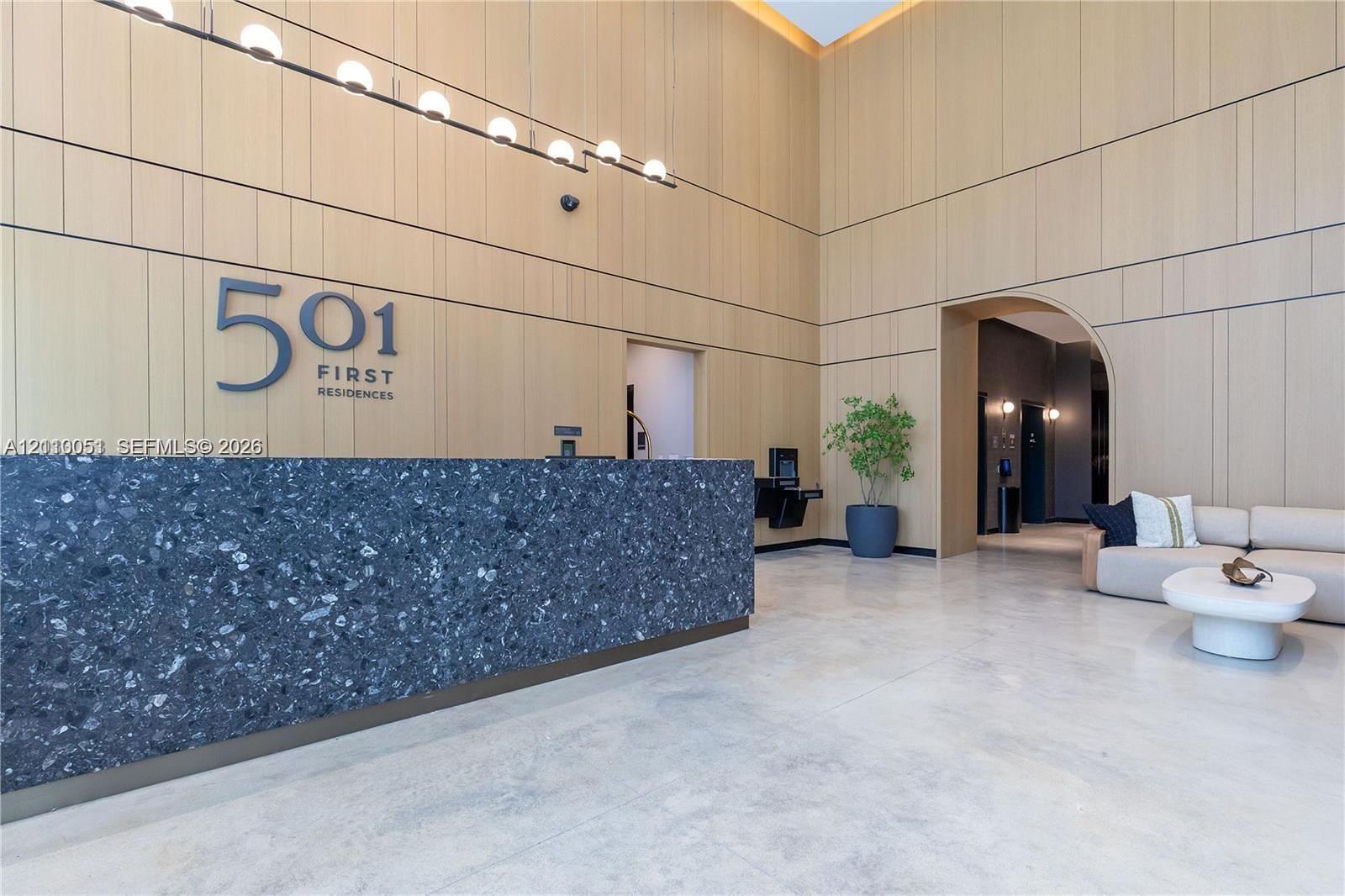 Lobby featuring double-height ceilings, wood-paneled walls, polished concrete flooring, a dark aggregate reception desk, and linear pendant lighting