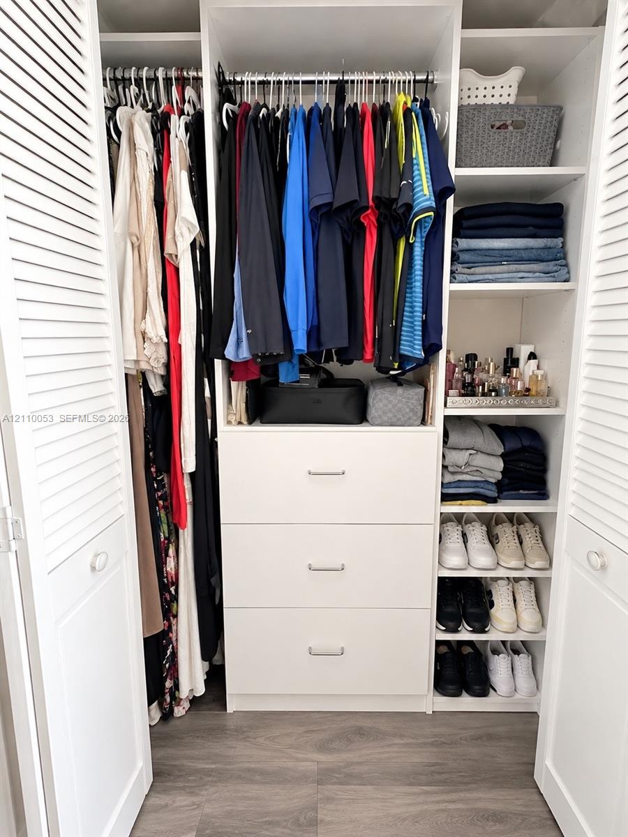 Closet featuring custom white built-in cabinetry, three soft-close storage drawers, adjustable open shelving, dual hanging rods, and white louvered bifold doors over wood-finish flooring
