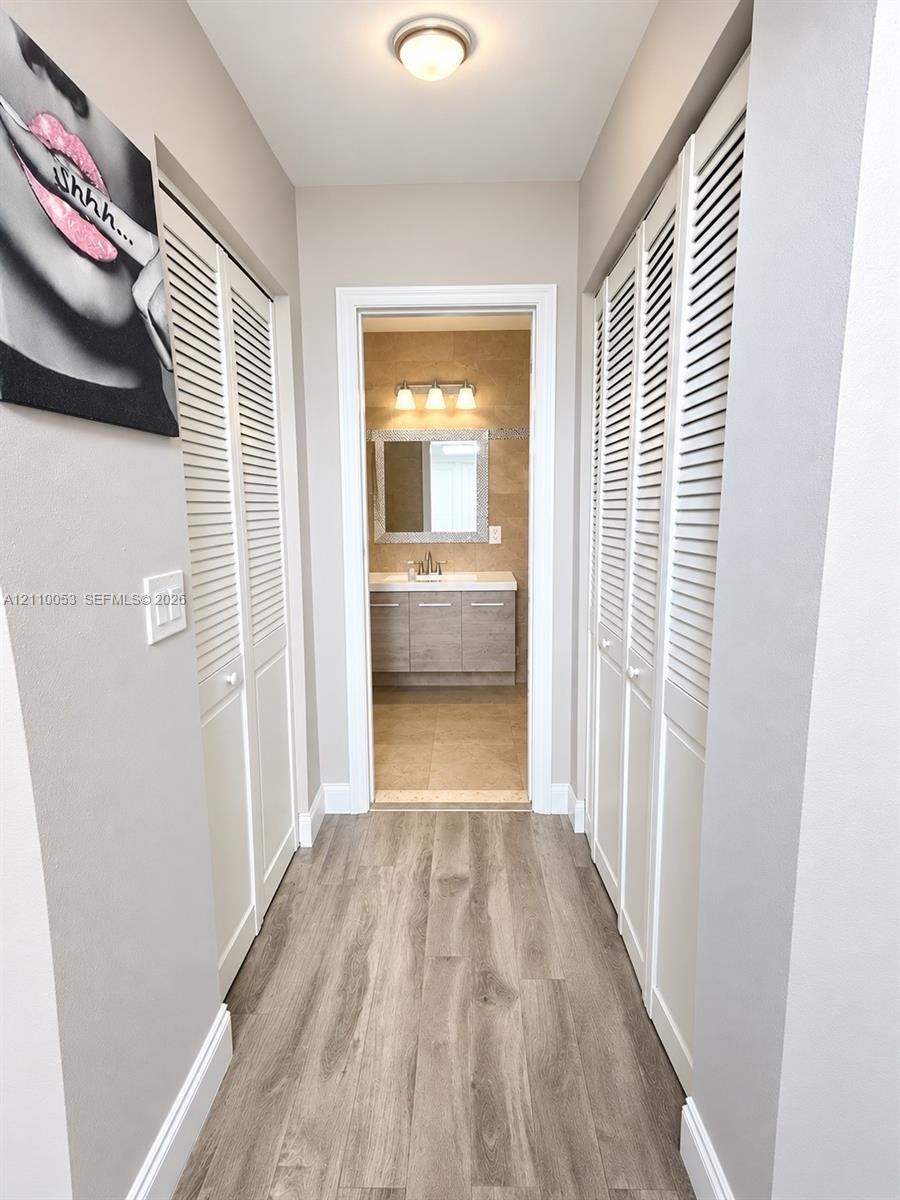 Hallway featuring dual sets of white louvered closet doors, wood-finish flooring, and a vanity area equipped with a mirror, three-light fixture, and stone-finish cabinetry