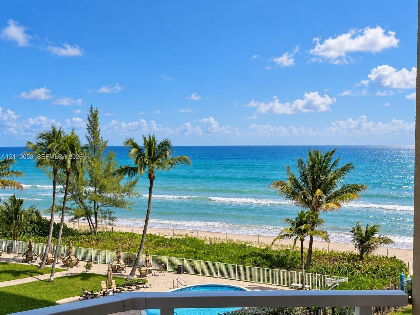 Private balcony overlooking a swimming pool, manicured lawn, sandy beach, and oceanfront horizon
