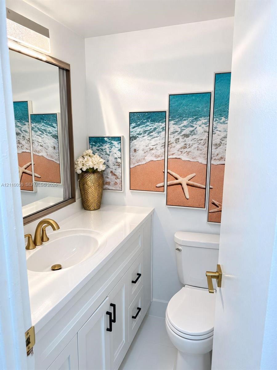 Vanity with white solid-surface countertop and undermount sink, brass-finish widespread faucet, white shaker-style cabinetry with matte black hardware, and rectangular vanity mirror with integrated light fixture