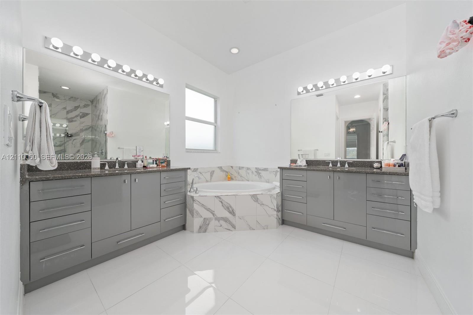 Dual vanities featuring gloss-finish cabinetry, stone countertops, integrated basins, and polished hardware, complemented by a tiled soaking tub and porcelain tile flooring